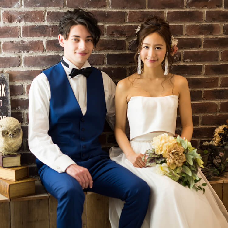 レンガ造りの壁の前でポーズをとった、白のウェディングドレスを着た花嫁と、青のベストを着た新郎